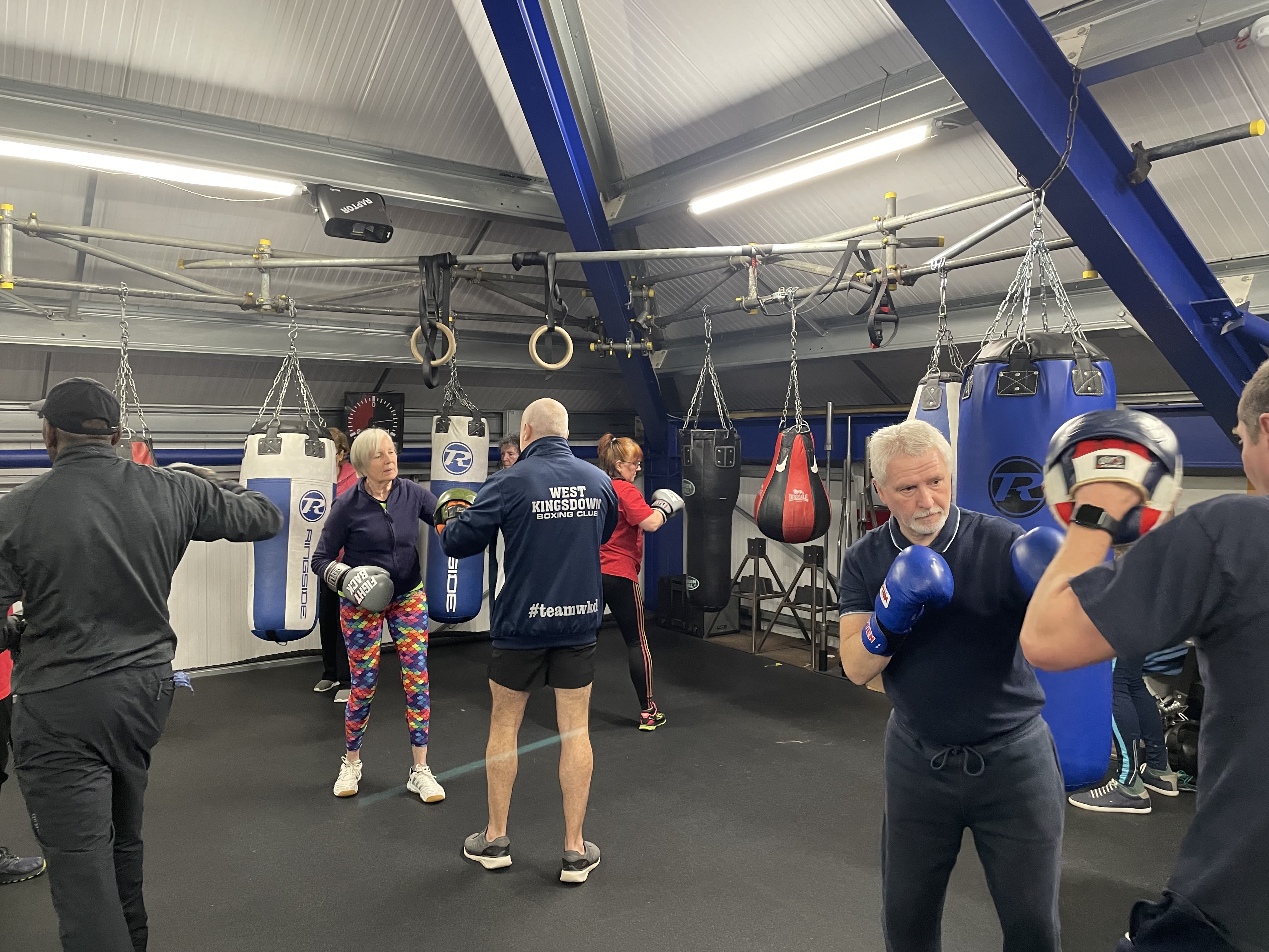 Coaches engage in individual pad work with different attendees living with Parkinson's, behind them, other goers practice on the punch bags.