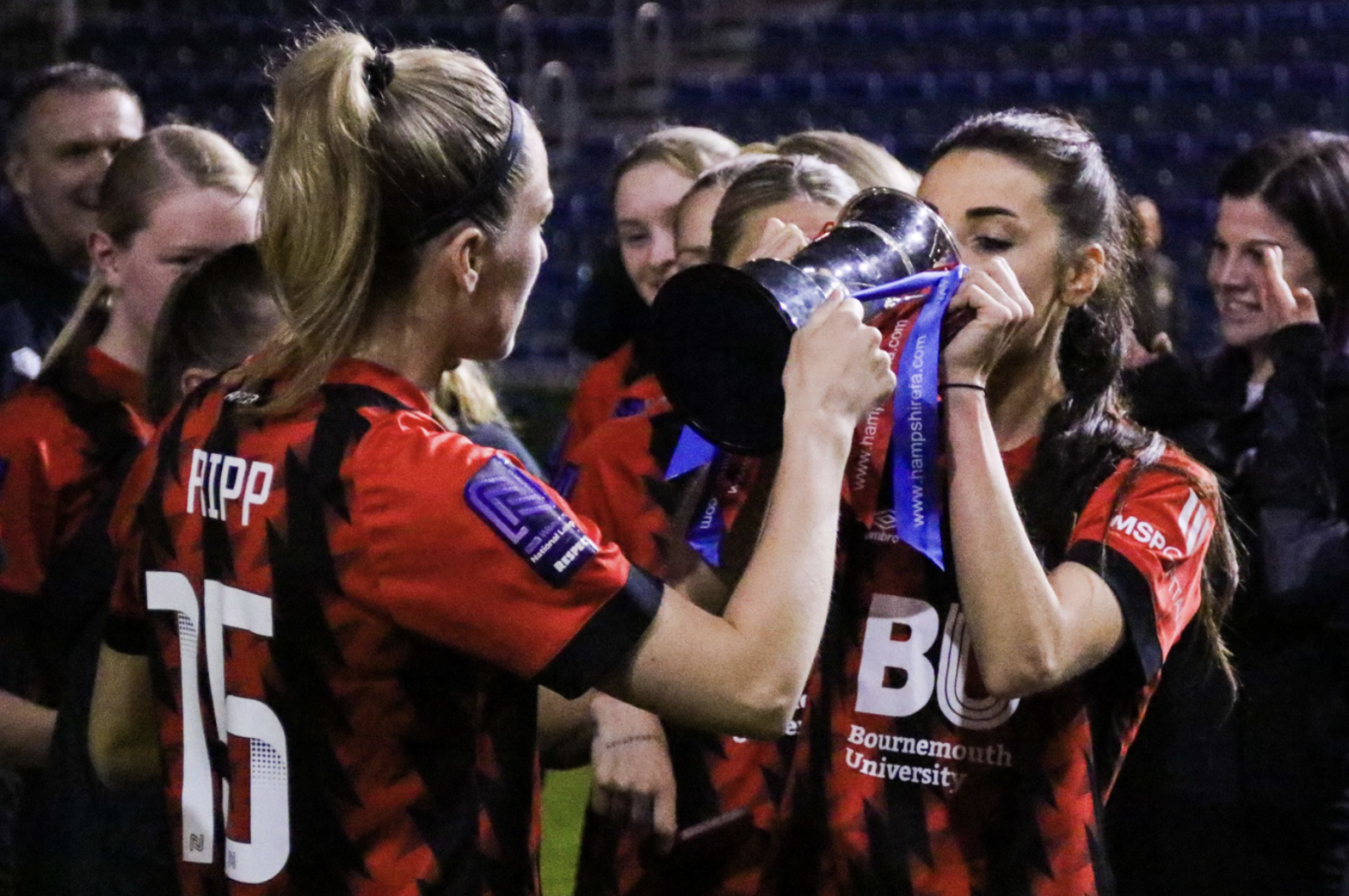 Kelly Fripp helping Maisy Smith drink out of the Hampshire FA Cup. They are both wearing the Bournemouth home shirt.