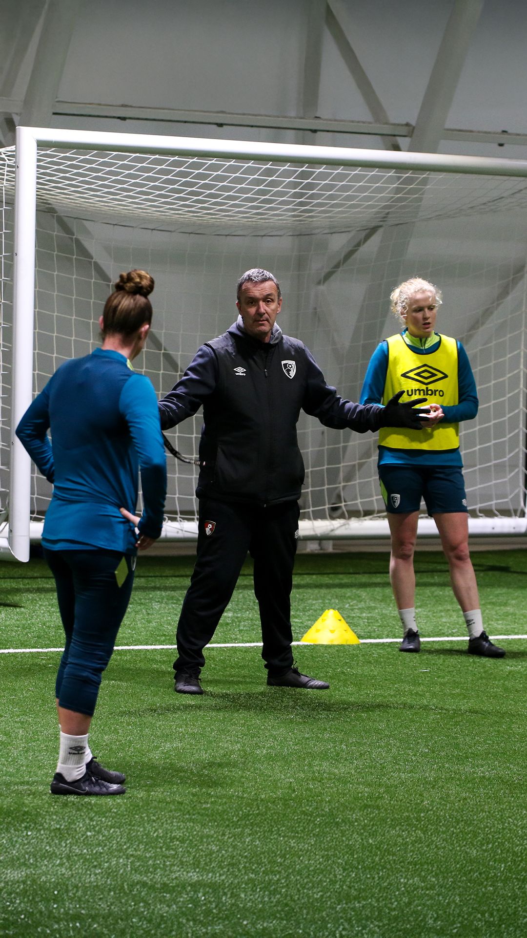 Steve Cuss with his arms spread out whilst speaking to Molly Barron-Clark. Amber Treweek is stood behind Steve Cuss wearing a yellow Umbro bib